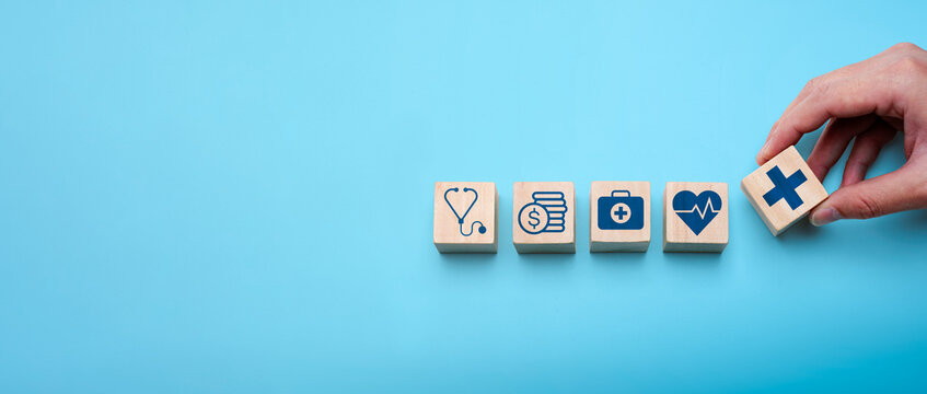 Healthcare planning and medical insurance concept with hand arranging wooden blocks showing icons of stethoscope, finance, first aid, heart health, and cross symbol on blue background