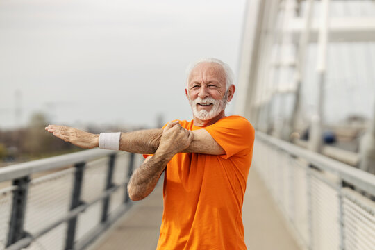 Portrait of an old sportsman in shape stretching his arms on a bridge. - Powered by Adobe