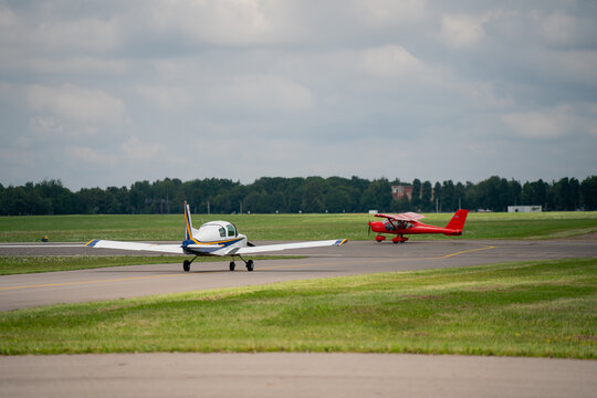 Two small single-engine airplanes taxiing on airport runway with grass field and trees in background captured from ground level view at general aviation airfield
