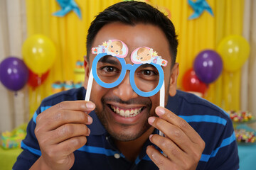 Happy man holding blue baby-themed party glasses over his eyes, smiling broadly at a lively celebration with colorful balloons. Joyful moment at a baby shower or birthday party.