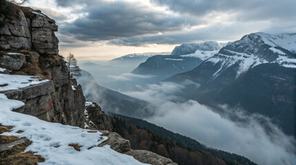 A dramatic view from a rocky ledge overlooking a mist-covered mountain valley, with snow-dusted peaks beyond. The sky is overcast, and the diffused light highlights the textures of the snow and rock. 