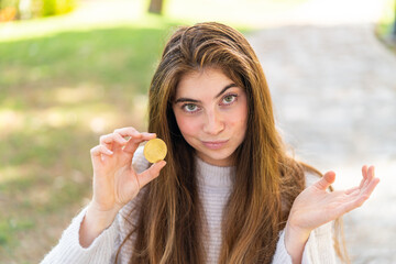 Young pretty caucasian woman holding a Bitcoin at outdoors making doubts gesture while lifting the shoulders