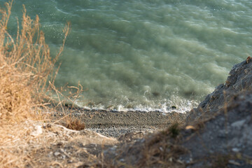View from a high cliff down to the turquoise sea and a narrow rocky beach, with dry grass and rugged rocks in the foreground.