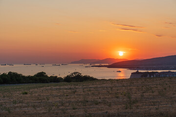 Sunset over the sea with ships on the water, mountains on the horizon, and a dry coastal field in the foreground. Gelendzhik