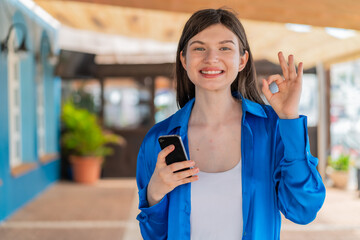 Young pretty woman at outdoors using mobile phone and doing OK sign