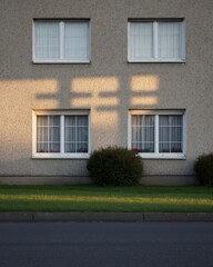 Sunlight casts long shadows on a light beige apartment building's windows.  Small plants are below the windows