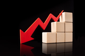 A red downward zigzag arrow next to with stacked wooden blocks, symbolizing a declining trend on a reflective black surface. High quality photo