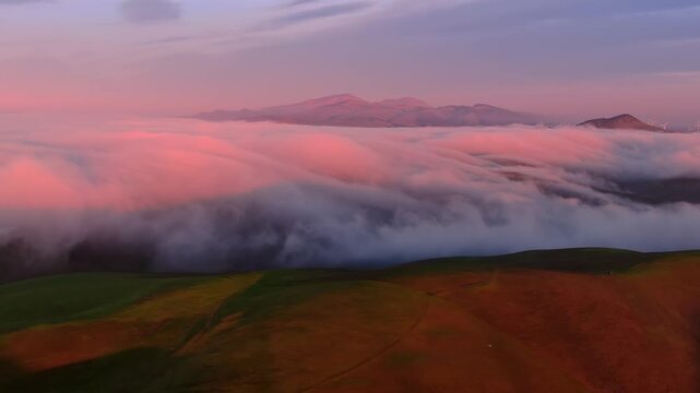 Fog rolls over the green hills of Altamont Pass, Livermore, California, USA, at sunset. The fog is illuminated by the pink light of the setting sun, creating a beautiful and ethereal scene.