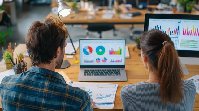 Team analyzes charts on laptops in office. Back view of man and woman studying data. For web, marketing, and editorial use.