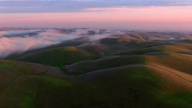 Aerial view of fog rolling over green hills at sunset in Altamont Pass, Livermore, California, USA. Mountains and wind turbines are visible in the distance.