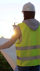 Woman an architect or engineer wearing safety hard hat and vest holding blueprint while inspecting...