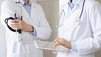 Two doctors wearing white coats and stethoscopes, using a digital tablet, discussing a patient's...