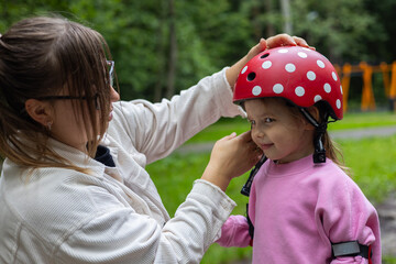 A young mother places a red helmet with white polka dots on her smiling daughter's head before...