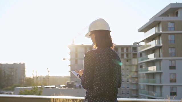 Female architect wearing a hard hat is taking notes with a tablet computer while inspecting a building construction site at sunrise in the morning