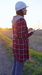 Woman engineer wearing a white protective helmet is taking notes with a clipboard in a field with...