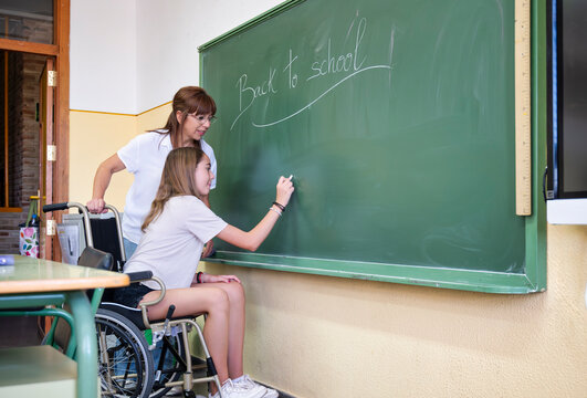 Schoolgirl in wheelchair writing on chalkboard with supportive teacher during inclusive classroom activity on first day of school