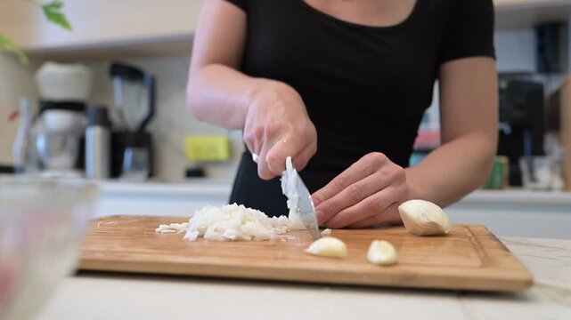 Close-up view of woman slicing fresh garlic and onion on a wooden cutting board in a stylish home kitchen. Perfect for food blogs, healthy recipes, or culinary visuals.