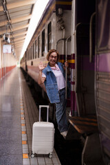 Asian older female traveler smiles waves boarding train station platform with suitcase embark...
