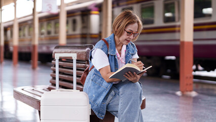 Asian senior female traveler writes journal notebook train station platform documenting journey planning trip waiting for adventure vacation concept © Pichsakul