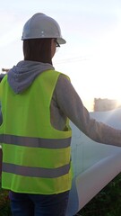 Female engineer wearing safety hard hat and vest holding blueprint while inspecting a construction...