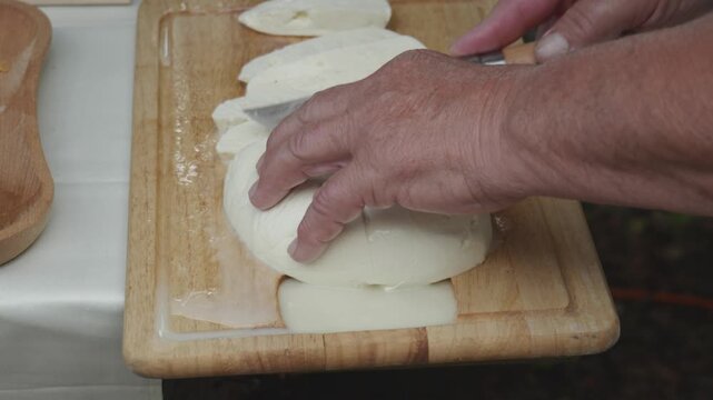 Close up of a man using knife to slice fresh traditional sheep cheese on a wooden cutting board