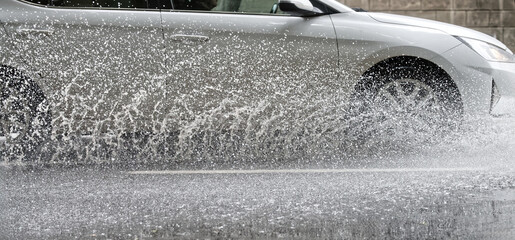 A white car drives through a large puddle, creating a dramatic splash of water from its wheels...