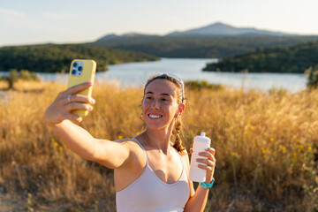 Young athletic woman taking a selfie with a yellow smartphone and holding a white water bottle in...