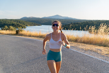 Muscular young woman smiling while running on a country road near a lake during summer