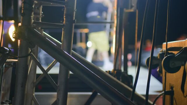 Cinematic close-up of concert stage metal structure at night, with blurred performer in background. Highlights technical setup, ideal for backstage, editorial, and music event themes.