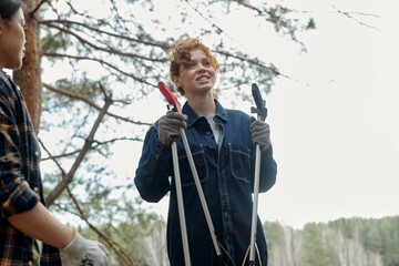 Young adult woman smiling while holding gardening shears outdoors, standing near another young...
