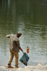 Young adult Black man cleaning riverbank using grabber tool and collecting plastic bottle and trash...