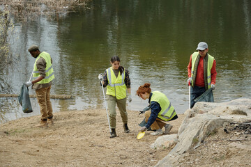 Group of young adult and middle aged multiethnic men and women cleaning lakeshore, wearing safety vests, collecting trash in bags, working together on environmental cleanup