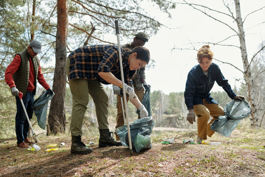 Group of young adult and middle aged multiethnic men and women collecting litter outdoors, picking up trash with gloves and bags in forest, participating in environmental cleanup activity