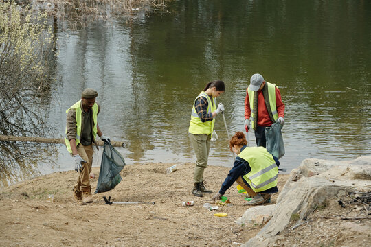 Group of young adult and middle aged multiethnic men and women cleaning up litter along lakeshore, wearing safety vests and gloves, collecting trash into large garbage bags