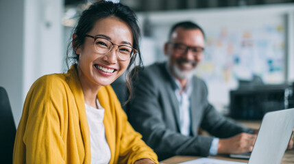 Smiling asian woman in yellow cardigan with man in suit at desk using laptop in office