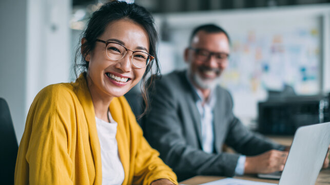 Smiling asian woman in yellow cardigan with man in suit at desk using laptop in office