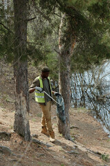 Obraz premium Black young adult man cleaning forested area near water using grabber tool and holding trash bag, wearing safety vest and gloves, participating in outdoor environmental cleanup