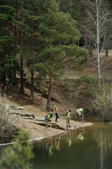 Group of middle aged men wearing safety vests working together near forest lake shore, performing environmental cleanup and collecting debris from water and ground