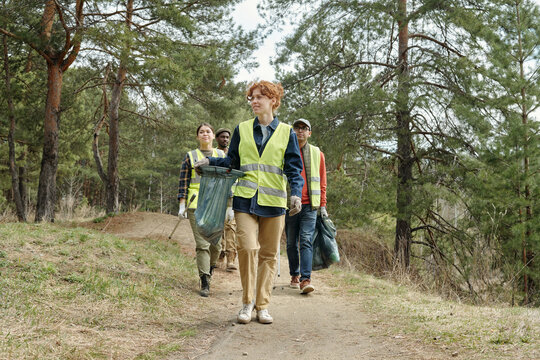 Group of men and women walking along forest path collecting trash wearing safety vests participating in outdoor community cleanup event