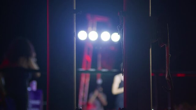 Side-stage backlit view framed by metal poles, showing stage lights and partial concert scene. Highlights industrial aesthetics and dynamic lighting of live events.