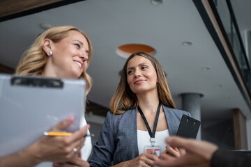 Businesswomen wearing lanyard and holding clipboard discussing in modern office building