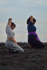 On warm sands, two teen girls sit in identical yoga poses, kneeling with arms positioned in...
