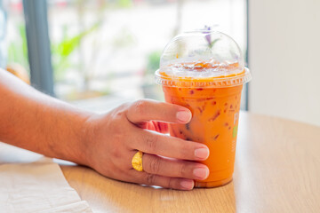 A hand holding a plastic cup of iced Thai milk tea on a wooden table in a bright cafe setting. Perfect for lifestyle, beverage, or takeaway drink concepts.