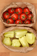 This bird's-eye view image shows two paper bags, one filled with red tomatoes and the other with light green bell peppers, placed on a wooden surface
