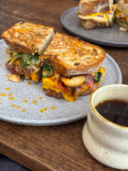 Delicious grilled sandwich with melted cheese, vegetables, and meat, served on a rustic ceramic plate with a cup of black coffee. Cozy cafe setting with wooden table and artisan presentation
