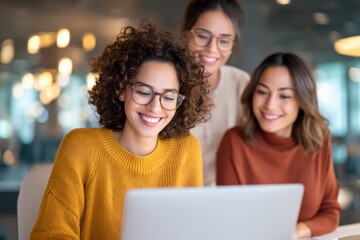 Three young women wearing glasses smiling and looking at a laptop in a bright, modern office setting.