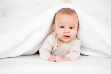 a baby, a smiling baby on a white bed under a blanket on his tummy at home, a healthy, joyful little baby boy in a beige bodysuit, a baby's sleep