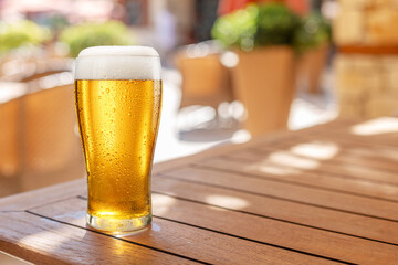 Glass of chilled beer on wooden table and blurred summer terrace at the background.