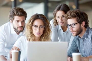 Four young professionals wearing glasses collaborate closely while looking intently at a laptop screen, with coffee cups nearby in a modern office setting.