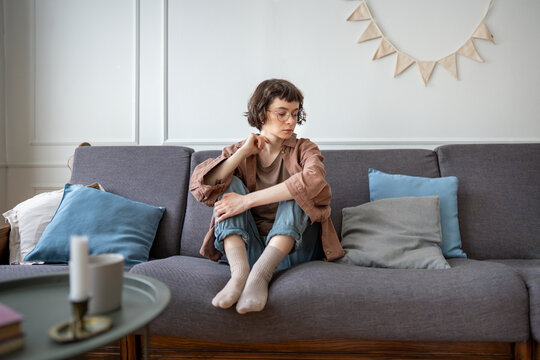 Sad tired teen girl in eyeglasses sits on couch lost in depressed thoughts needs psychologies support. Upset pensive young woman frustrated thinking about communication problems feels solitude, empty.